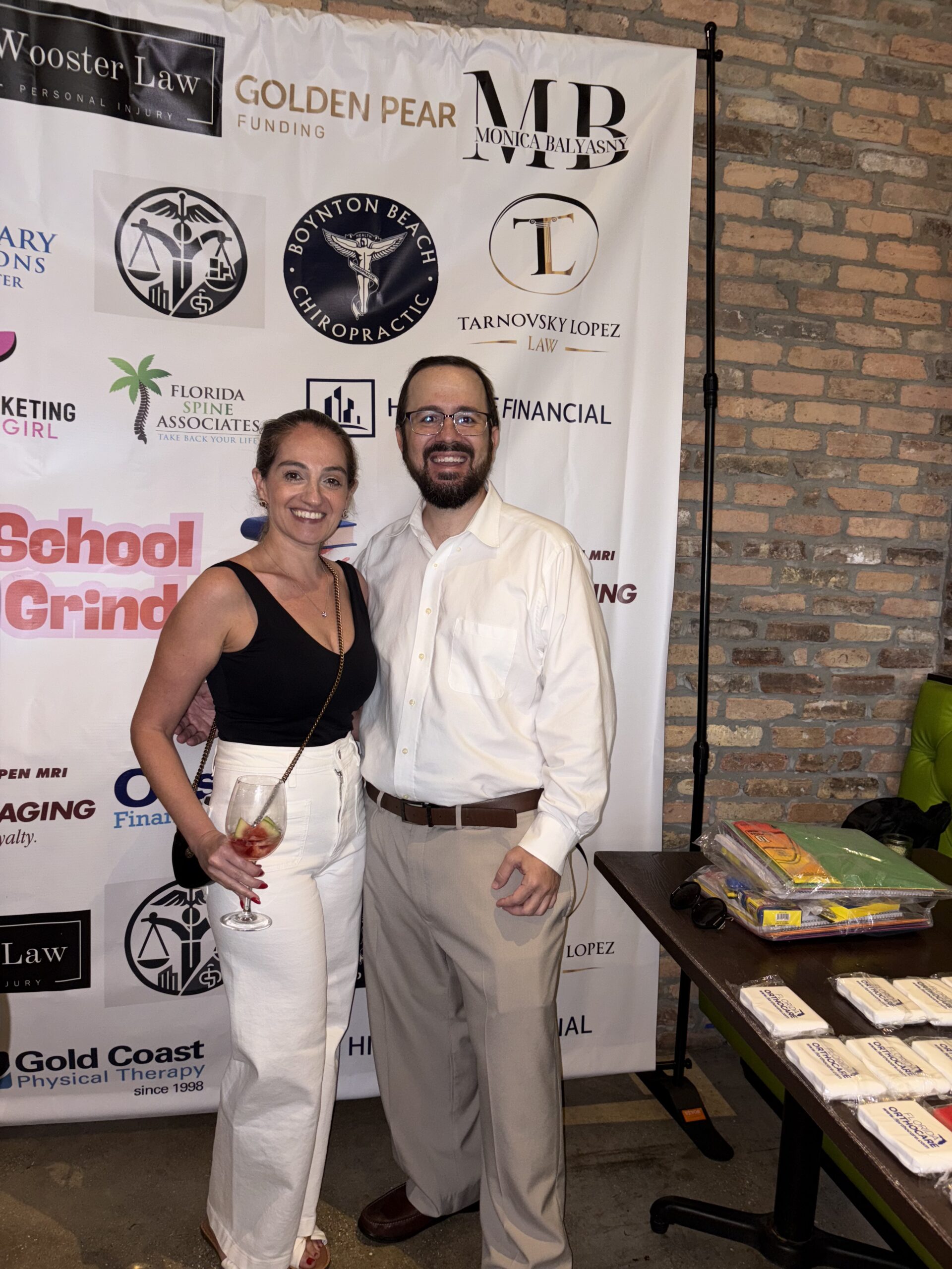Irina Tarnovsky and Aex Lopez of Tarnovsky Lopez Law at Windsor Imaging’s Back2School Back2Grind event, standing in front of a sponsor backdrop featuring the firm’s logo among other local businesses.