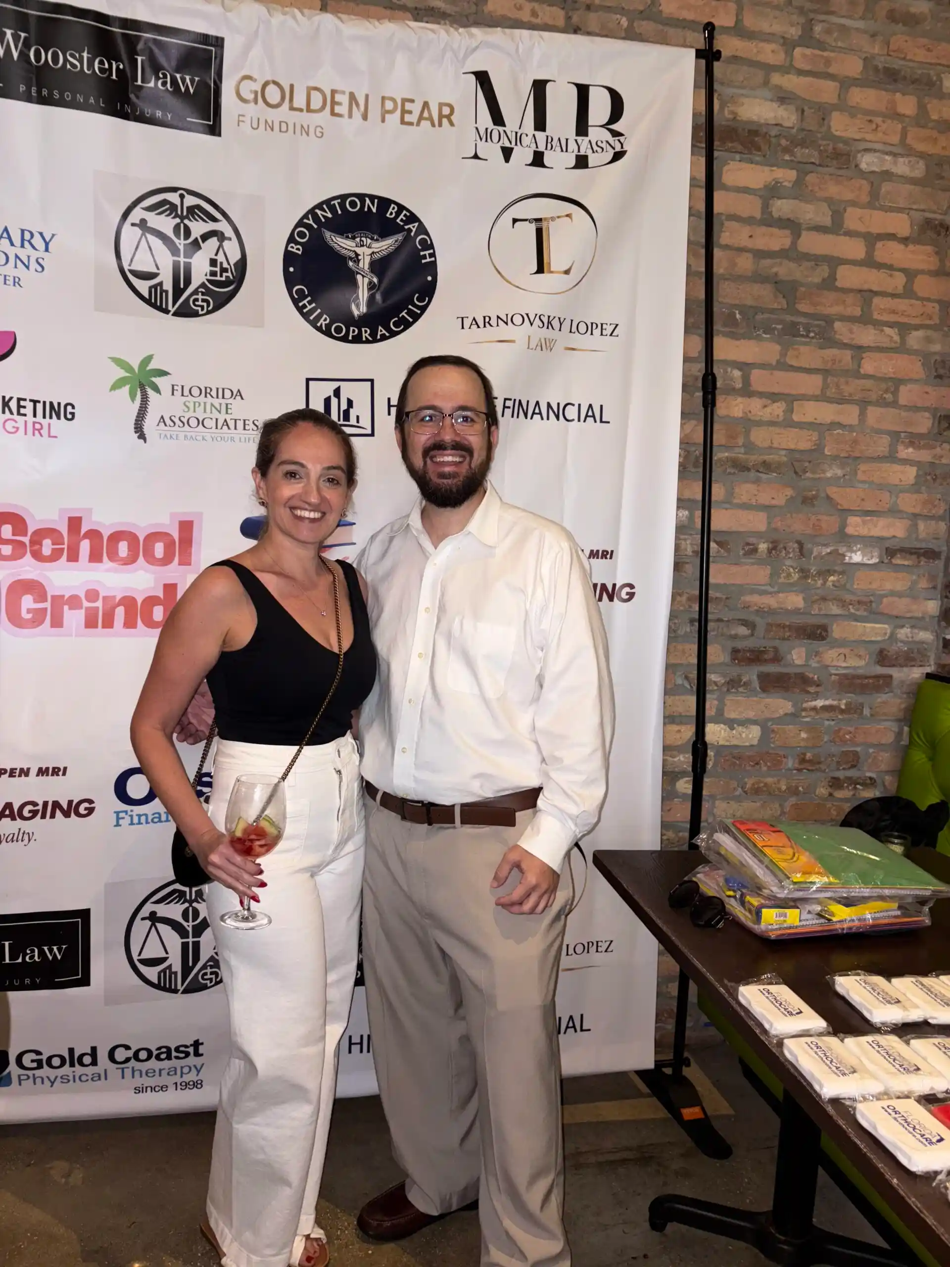 Irina Tarnovsky and Aex Lopez of Tarnovsky Lopez Law at Windsor Imaging’s Back2School Back2Grind event, standing in front of a sponsor backdrop featuring the firm’s logo among other local businesses.